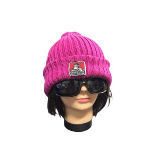 May include: A bright pink ribbed knit beanie with a Ben Davis logo patch. The beanie is worn with black sunglasses on a mannequin head. The logo patch features a man in overalls.