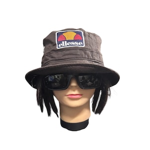 May include: A brown bucket hat with a square patch featuring the brand name "ellesse" in white, with a red, yellow, and blue logo. The hat is worn by a mannequin wearing black sunglasses.