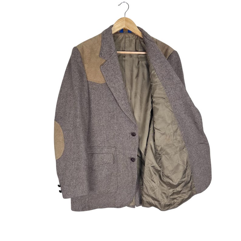 May include: A brown herringbone blazer featuring tan suede shoulder patches and matching elbow patches. The jacket has a two-button front closure, two front flap pockets, and a solid tan interior lining. It is displayed on a wooden hanger.