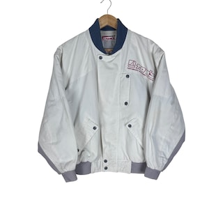 May include: A white bomber jacket featuring a dark blue ribbed collar and grey ribbed cuffs. The jacket has a front zip closure with snap buttons, two front pockets, and the red and white TOM'S logo embroidered on the upper left chest area.