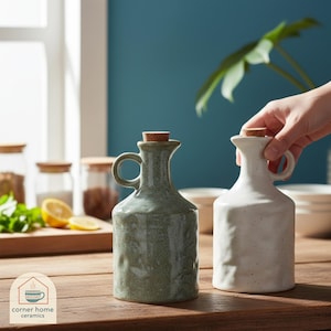 May include: Two ceramic oil bottles with cork stoppers. One is a speckled sage green, the other is off-white. Both have a handle and a spout. The bottles are on a wooden surface. The logo for "corner home ceramics" is in the lower left corner.