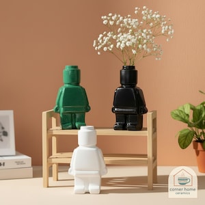 Może przedstawiać: Trzy ceramiczne wazony w kształcie figurek, zielone, czarne i białe, są wystawione na małej drewnianej półce. Czarny wazon zawiera białe kwiaty. Wazony mają błyszczące wykończenie i są ustawione na brzoskwiniowym tle.