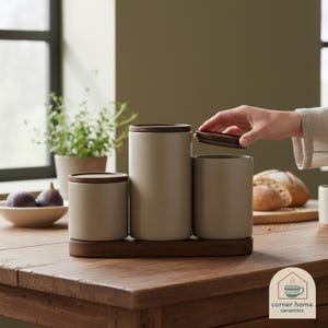 May include: A trio of beige ceramic storage jars with dark brown wooden lids, presented on a matching wooden tray. The image shows the jars in a well-lit room. The jars are of different heights. The table is made of wood.