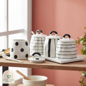 Puede incluir: Un conjunto de botes de cocina de cerámica blanca y una taza con detalles negros. Los botes presentan diferentes diseños, incluyendo lunares, rayas y un diseño de cuadrícula. El conjunto incluye una bandeja y cucharas.