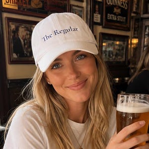 May include: A white baseball cap with the text The Regular embroidered in navy blue on the front. The cap features a curved brim and a structured crown, constructed from a cotton-like fabric with a button detail at the top center of the headwear.
