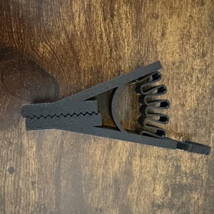 May include: A black, foam-like capo for a stringed instrument. The capo has a hinged design with a serrated clamp and a series of curved, finger-like grips. The background is a wooden surface.
