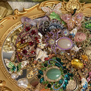 Könnte beinhalten: Eine Sammlung verzierter Broschen und Anstecknadeln, darunter Schmetterlings- und Katzenmotive, auf einem goldgerahmten Spiegel. Die Broschen sind mit farbigen Edelsteinen und Kristallen in Rot-, Grün-, Gelb- und Lilatönen besetzt. Das Gesamtthema ist Vintage und glamourös.