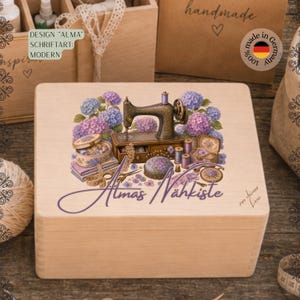 Könnte beinhalten: Eine helle Holzbox mit einem aufgedruckten Motiv einer alten Nähmaschine, umgeben von violetten und blauen Hortensien, Garnrollen, einem Nadelkissen und einer Schere, ergänzt durch den violetten Schriftzug Almas Nähkiste auf der Vorderseite.