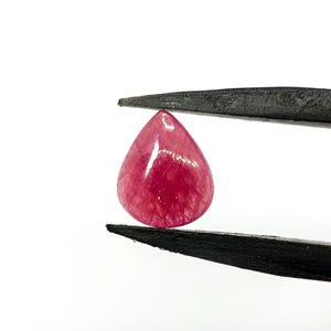 May include: A pear-shaped, translucent gemstone with a deep magenta-pink hue and visible internal inclusions. The stone is held securely between the two pointed tips of a pair of black metal tweezers against a plain white background.