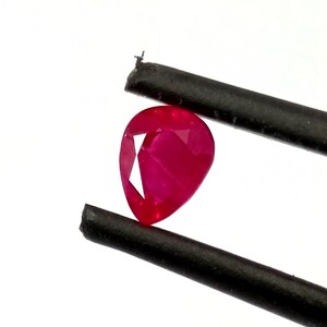 Peut inclure: Une pierre précieuse rubis taillée en forme de poire, d'une couleur rouge profond, maintenue entre deux pinces en métal noir. La pierre présente des reflets lumineux internes et une surface polie, mettant en valeur sa taille précise.