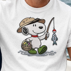 May include: White t-shirt featuring a cartoon Snoopy character fishing. Snoopy is wearing a brown hat, green boots, and a red collar, holding a fishing rod with a fish and a basket of Easter eggs.