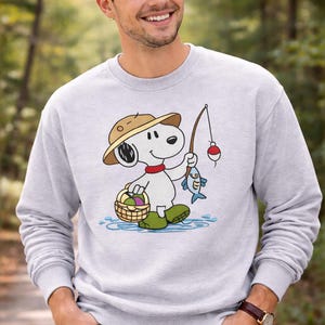 May include: Light gray sweatshirt featuring a cartoon Snoopy character fishing. Snoopy is wearing a brown hat, red collar, and green boots, holding a fishing rod with a fish and a basket of eggs. The design is set against a water background.
