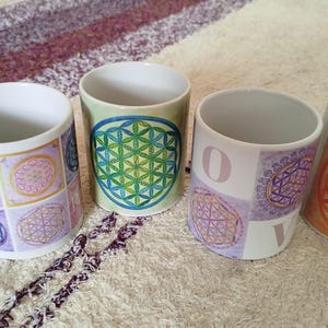 May include: Four white ceramic mugs with colourful geometric designs. One mug has a handle and the word "amour" on it. The other mugs feature the "Flower of Life" pattern in various colours, including green, purple, and orange. The mugs are on a patterned surface.