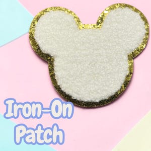May include: A white, textured, fuzzy, sherpa-like fabric patch in the shape of a mouse head silhouette. The patch features a thick, shimmering gold glitter border that outlines the entire shape. The text Iron-On Patch is visible.