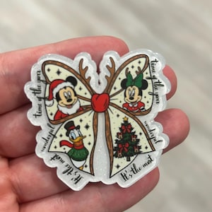 Könnte beinhalten: Eine schleifenförmige Acryl-Anstecknadel mit Glitzer-Finish, die Mickey Mouse mit Weihnachtsmütze, Minnie Mouse mit grüner Schleife, Donald Duck mit Winterschal und einen Weihnachtsbaum zeigt. Der Text lautet, It's the most wonderful time of the year.