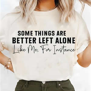 Op de afbeelding: Crèmekleurig T-shirt met de zwarte tekst "SOME THINGS ARE BETTER LEFT ALONE Like Me, For Instance". De persoon draagt het shirt, met een gouden ketting en armband zichtbaar.
