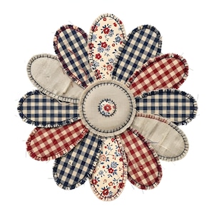 May include: A handcrafted, flower-shaped item with a patchwork design. The petals feature red and navy gingham, floral patterns, and solid beige fabric. The center is beige with a small floral design. The edges are stitched with dark thread.