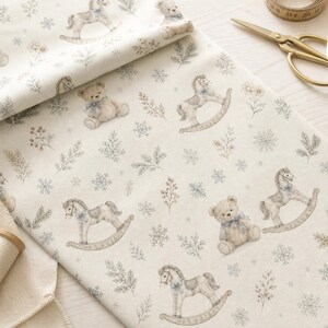 May include: A cream-coloured fabric featuring a repeating pattern of light brown teddy bears with blue bows, rocking horses, snowflakes, and sprigs of greenery. The textile is displayed with gold-toned scissors and a measuring tape nearby.