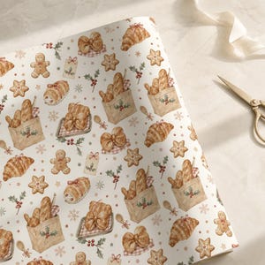 May include: A roll of wrapping paper featuring a repeating watercolor pattern of gingerbread people, croissants, baguettes in paper bags, star-shaped cookies, holly berries, and snowflakes on a white background, accompanied by gold scissors.
