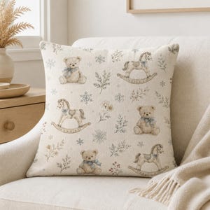 May include: A square throw pillow with a cream-coloured fabric base featuring a repeating pattern of light brown teddy bears with blue bows, wooden rocking horses, blue snowflakes, and delicate sprigs of winter foliage and small flowers.