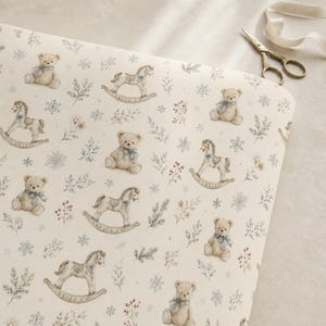 May include: A cream-coloured fabric featuring a repeating pattern of light brown teddy bears with blue bows, rocking horses, snowflakes, and sprigs of foliage. A pair of metal scissors with a ribbon is positioned near the top right corner.