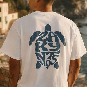 Könnte beinhalten: Ein weißes kurzärmeliges T-Shirt mit einer großen dunkelblauen Grafik auf der Rückseite. Die Grafik hat die Form einer Meeresschildkröte und enthält den Text ZAKYNTHOS in einer stilisierten, fetten Schrift, die das Innere der Schildkröte ausfüllt.