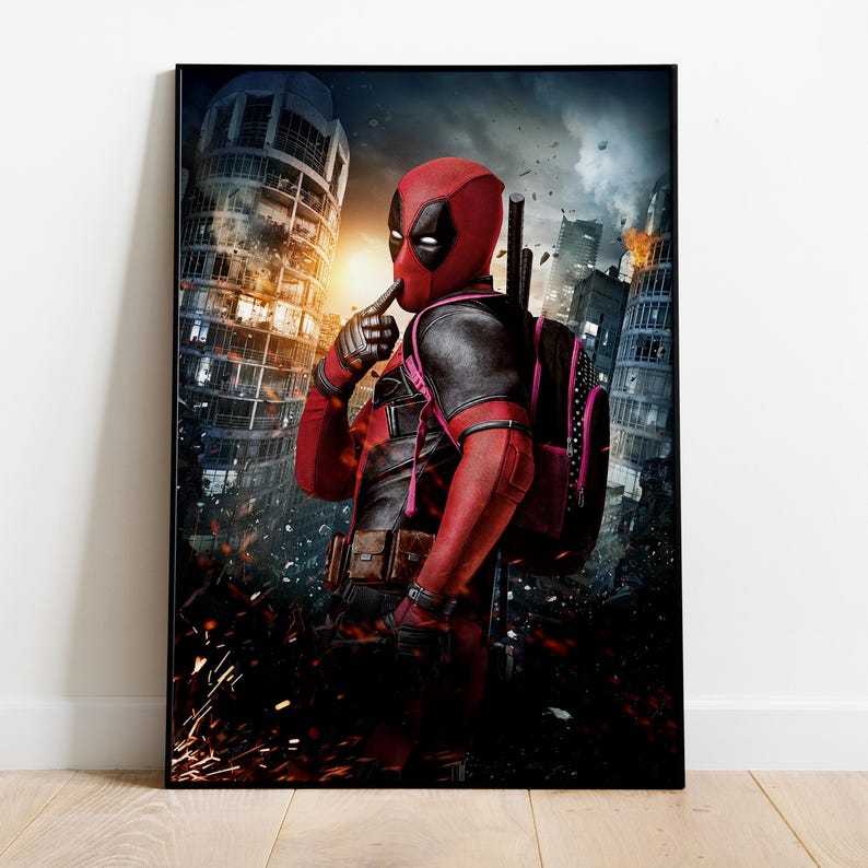 May include: A framed poster featuring the character Deadpool in his signature red and black costume, with a mask covering his face. He is in a city setting with a backpack and weapons, with a finger to his lips. The background shows a city in flames.