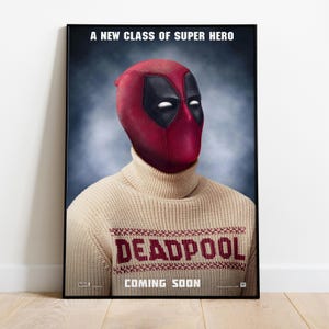 May include: A framed poster featuring Deadpool in a red and black mask and a beige turtleneck sweater. The sweater has the word "DEADPOOL" knitted across the chest. The poster includes the text "A NEW CLASS OF SUPER HERO" and "COMING SOON".