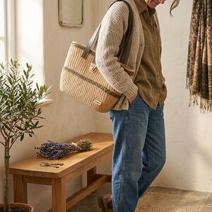 May include: A woven straw tote bag with leather handles is carried by a person. The bag has a rectangular shape with a dark stripe detail. The person is wearing a light-colored cardigan, a button-down shirt, and blue jeans. A wooden bench and a small potted plant are in the background.