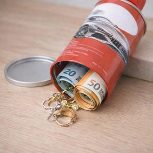 Op de afbeelding: Een rood-witte blik met een rode dop, hergebruikt als verborgen opbergcontainer. Opgerolde eurobiljetten en sieraden komen uit de opening. De blik heeft een autografiek.