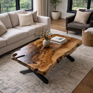 Peut inclure: Une table basse en bois avec des bords naturels, présentant un veinage naturel. La table a une forme irrégulière et repose sur des pieds en métal noir. Un vase et un livre sont posés sur la table.