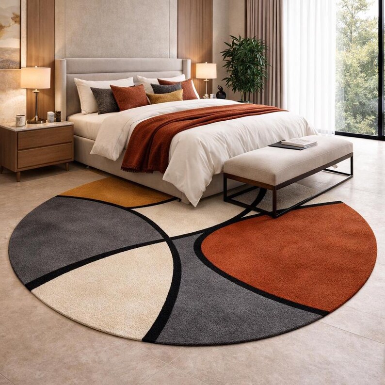May include: A round area rug with a geometric design in a bedroom setting. The rug features sections of gray, cream, orange, and yellow, outlined with black lines. The rug is placed in front of a bed with white bedding and a rust-colored throw.