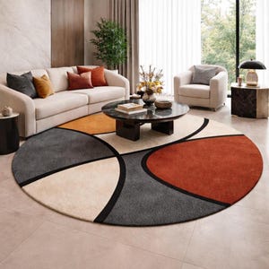 May include: A round area rug with a geometric design in shades of gray, orange, beige, and rust. The rug features a black border outlining the shapes. The rug is placed in a living room setting with a sofa, armchair, and coffee table.