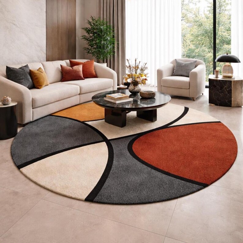 May include: A round area rug with a geometric design in shades of gray, orange, beige, and rust. The rug features a black border outlining the shapes. The rug is placed in a living room setting with a sofa, armchair, and coffee table.