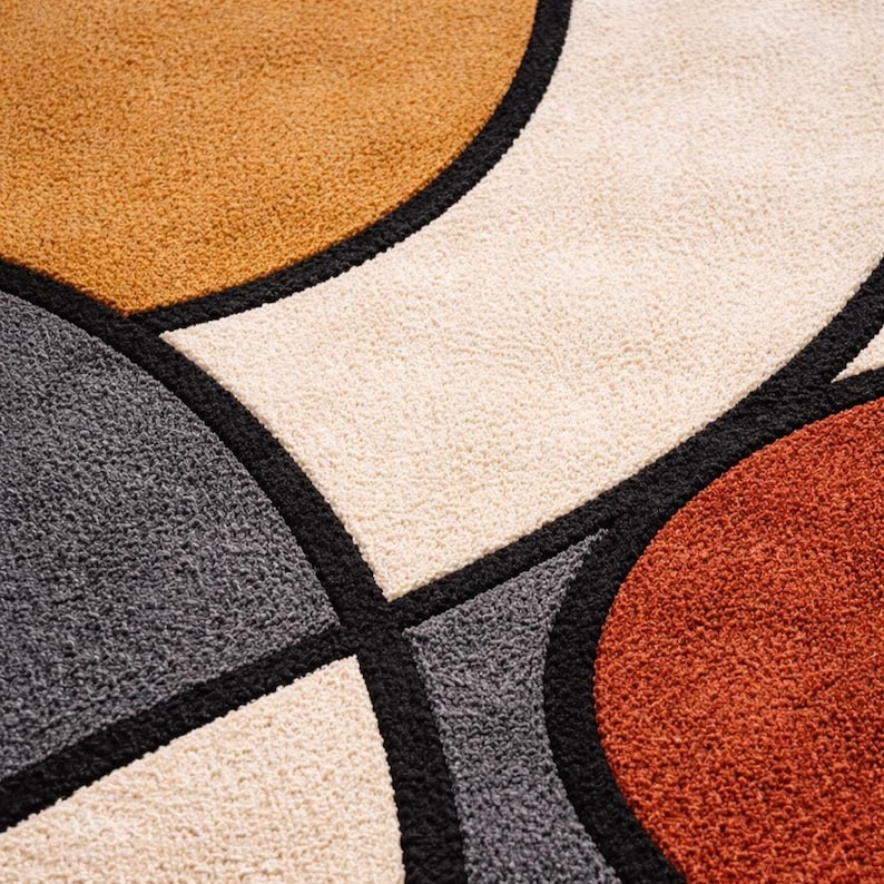 May include: Close-up of a geometric rug with a modern design. The rug features large, rounded shapes in shades of tan, cream, gray, and rust, outlined in black. The textured surface adds depth and visual interest to the abstract pattern.