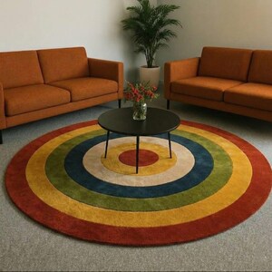 May include: A round rug with a rainbow design in shades of red, orange, yellow, green, blue, and cream. The rug is centered in a room with two orange sofas and a black coffee table. A potted plant is in the background.