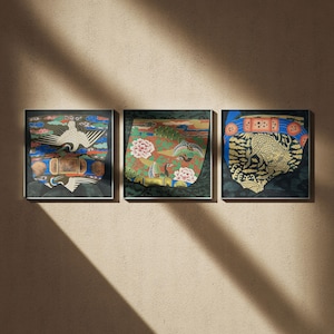 Op de afbeelding: Drie ingelijste kunstwerken geïnspireerd op Azië. Elk heeft een uniek ontwerp met levendige kleuren, waaronder een witte kraanvogel, roze bloemen en een gouden vis. Het kunstwerk staat tegen een donkergroene achtergrond, met zwarte lijsten.