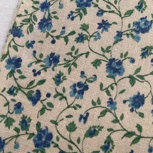 May include: A piece of woven fabric featuring a light beige background with a repeating pattern of small blue flowers and thin green vines. The textile has a visible cross-hatched weave texture throughout, showcasing a consistent floral print.