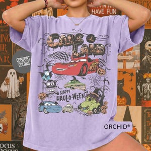 May include: A light purple t-shirt featuring a graphic print with the text "CARS LAND" and "HAPPY HAUL-O-WEEN". The design includes a red race car, a purple tow truck, a blue car, and a green car, all decorated with Halloween-themed elements.