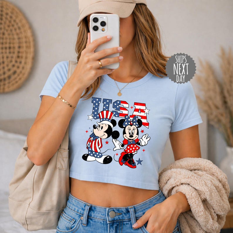 A woman is taking a selfie with a smartphone, wearing a blue crop top with an image of Minnie Mouse and Mickey Mouse, and holding a beige sweater.