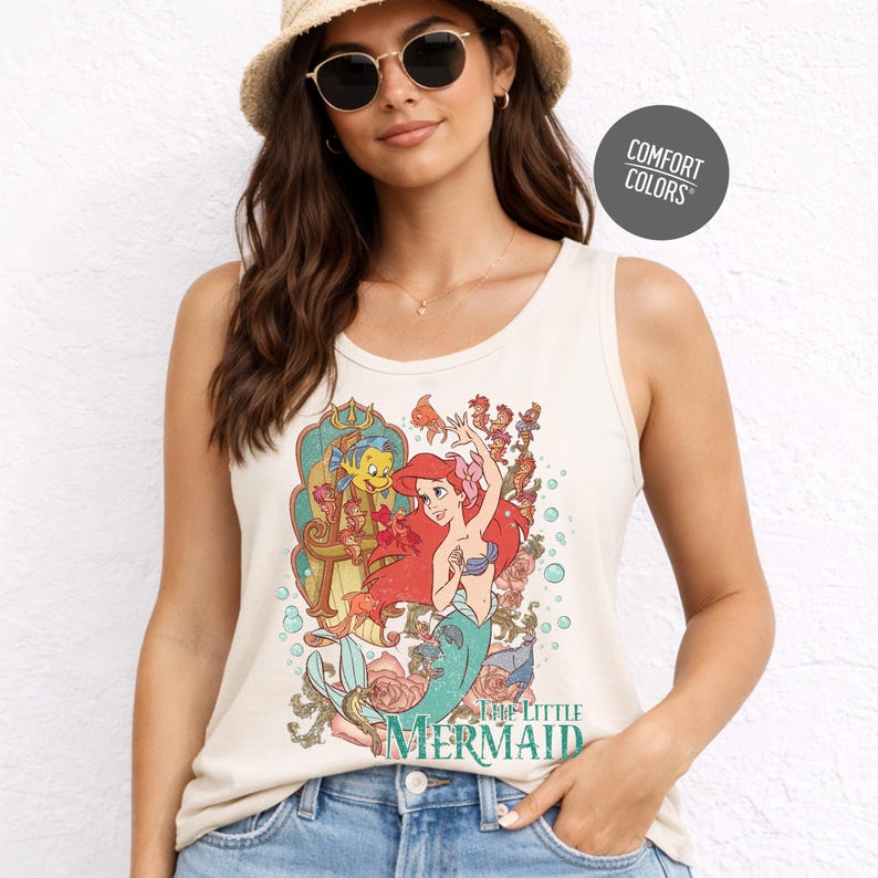A woman wearing a white tank top with a printed design of the Disney character Ariel from The Little Mermaid, smiling and posing for the camera.