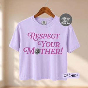 May include: A light purple short-sleeve t-shirt with a boxy fit. The front features the dark pink text "RESPECT YOUR MOTHER!" with a small silver disco ball replacing the letter "O". A circular tag reads "COMFORT COLORS BOXY FIT" and a label says "ORCHID".