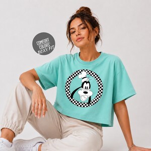 A woman wearing a turquoise crop top with a cartoon character graphic on it, posing against a white background.