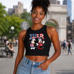 A young woman wearing a black crop top with an American flag design and Minnie Mouse on it, standing in front of a city street with a monument in the background.