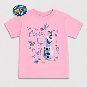 A pink t-shirt with a graphic design featuring the character Olaf from the Disney movie Frozen. The design includes Olaf holding a snowman and the text "Never Too Cool" written above him.
