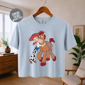 A light blue t-shirt with a cartoon image of a girl and a horse, along with a wooden hanger.