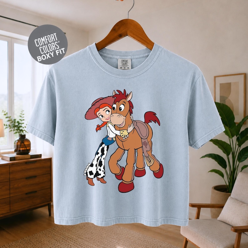 A light blue t-shirt with a cartoon image of a girl and a horse, along with a wooden hanger.
