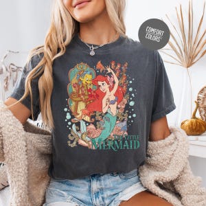 A woman wearing a gray t-shirt with a graphic of the mermaid character from the Disney film "The Little Mermaid".