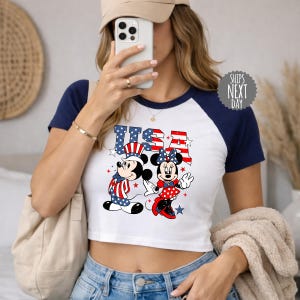 A woman is taking a selfie with a smartphone, wearing a white and blue crop top with a graphic of Minnie Mouse and an American flag design.