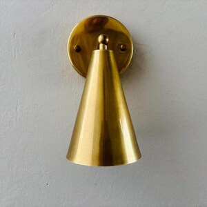 May include: A brass wall sconce with a conical shade and a circular backplate. The sconce has a polished finish and is mounted on a light-colored wall. The design is minimalist and modern, suitable for interior lighting.